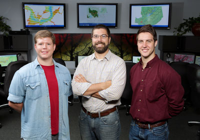 UAH seniors Casey Calamaio and Kel Markert flank their advisor Dr. Rob Griffin in the NASA SERVIR Earth Science Student Research lab at the National Space Science and Technology Center on campus. Behind them on the monitors is a vegetation index of Guntersville Lake, a sub-watershed analysis map around Guntersville and a map showing their study area.