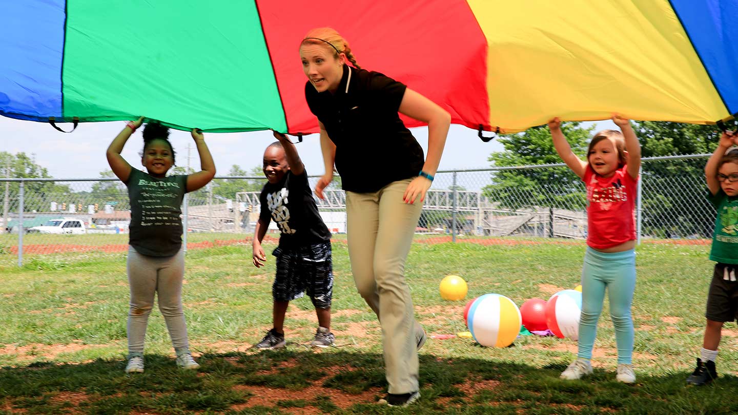 people playing the parachute game. The teacher is under a parachute while children lift the parachute for her to run under.