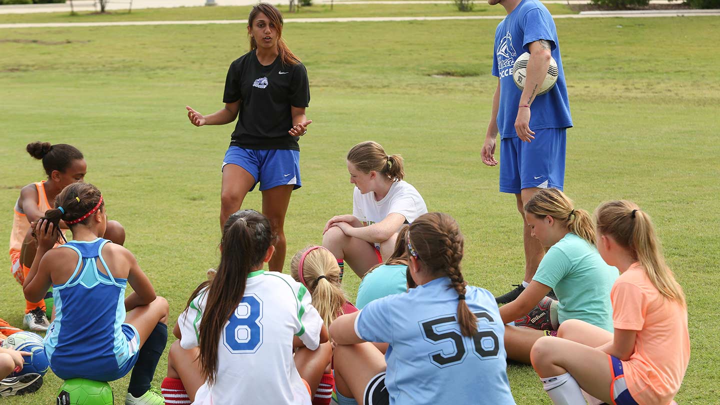 A soccer coach stands and speaks to a group of girls. The girls are setting around their coach, listening to her, setting on a soccer field.