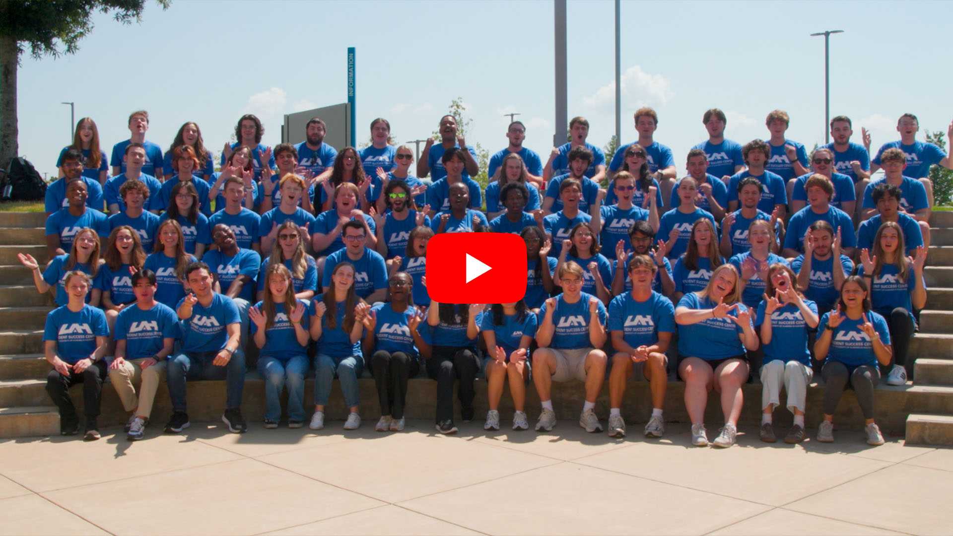 Photo of a large group on the UAH campus, smiling and holding up UAH spirit hand signs. A red play button indicates the image links to a video.
