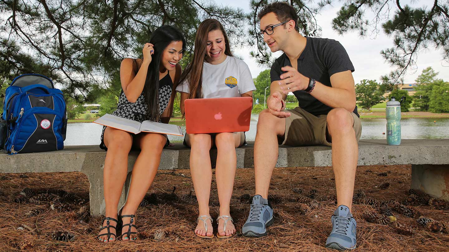 Three students setting on bench, while looking at a laptop screen. Students smiling, laughing.