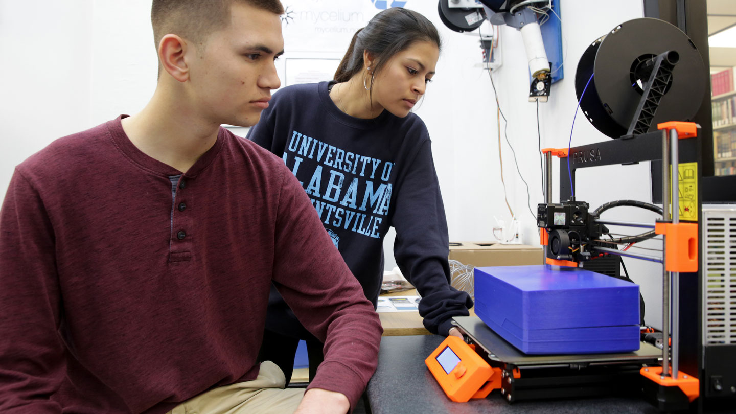 Two U A H students use the 3 D printer in the library's makerspace lab.
