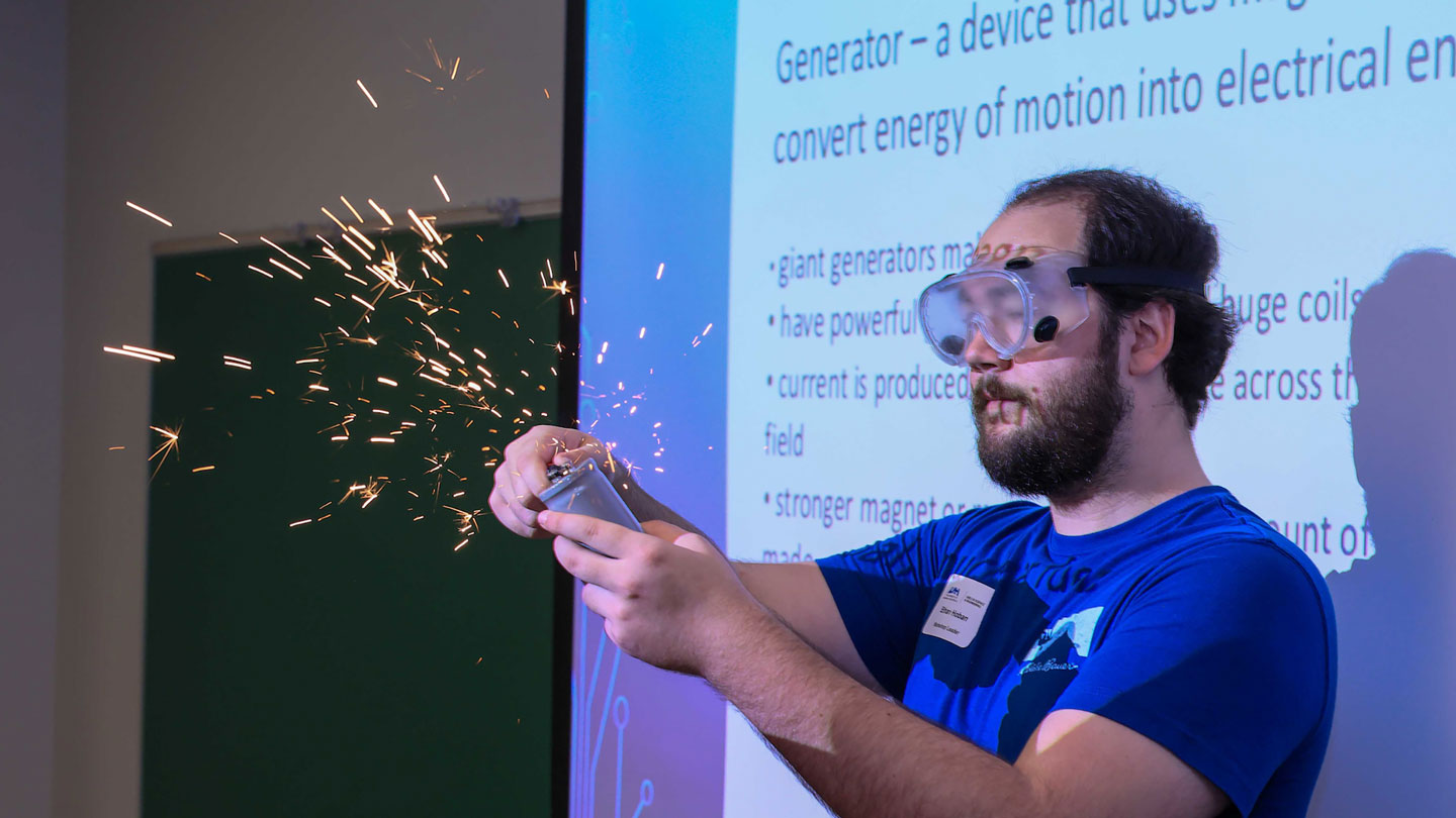 A man wearing safety goggles gives a presentation and uses sparks as a visual aid