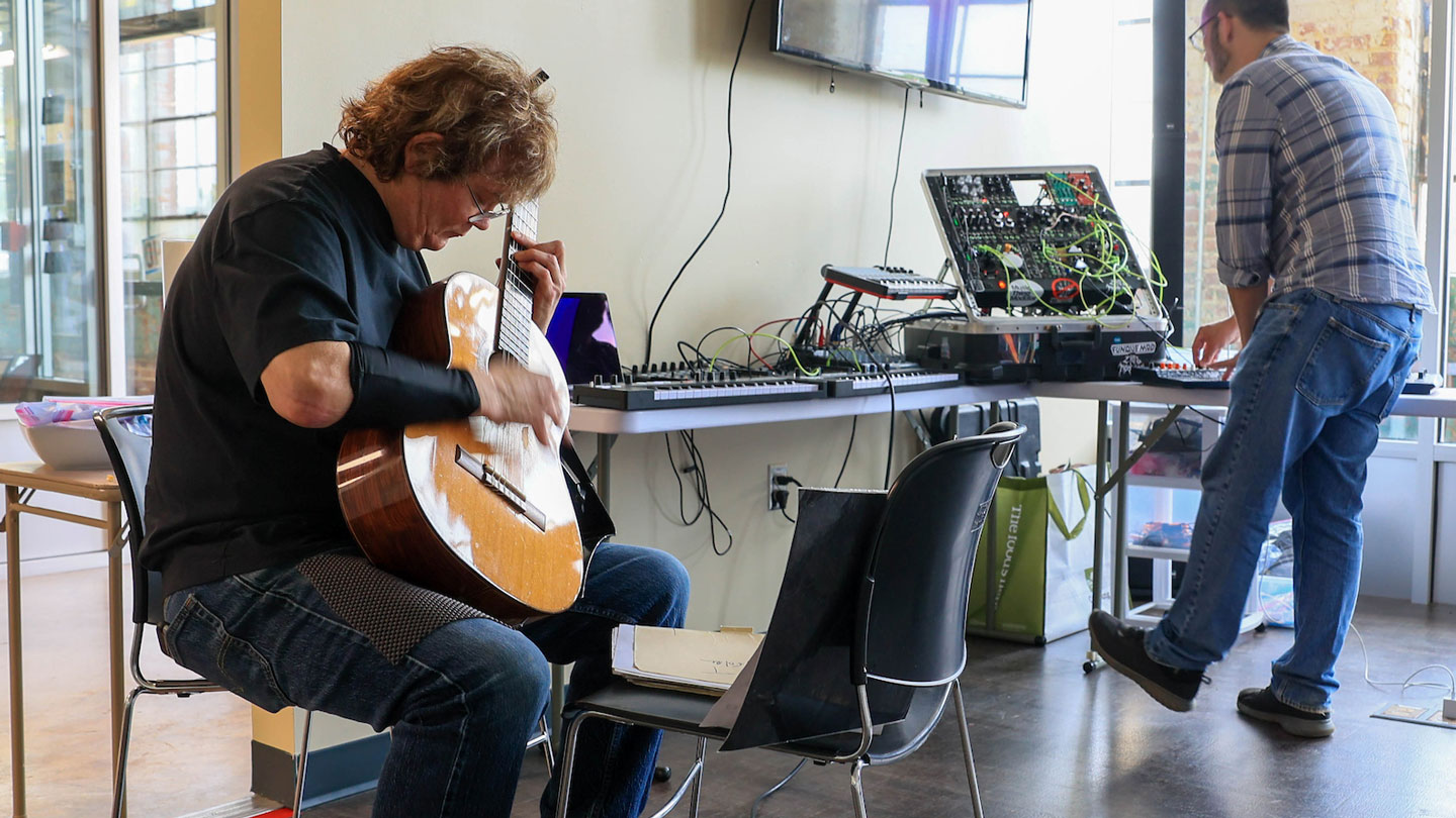 A man plays the guitar while some else fixes sound equipment