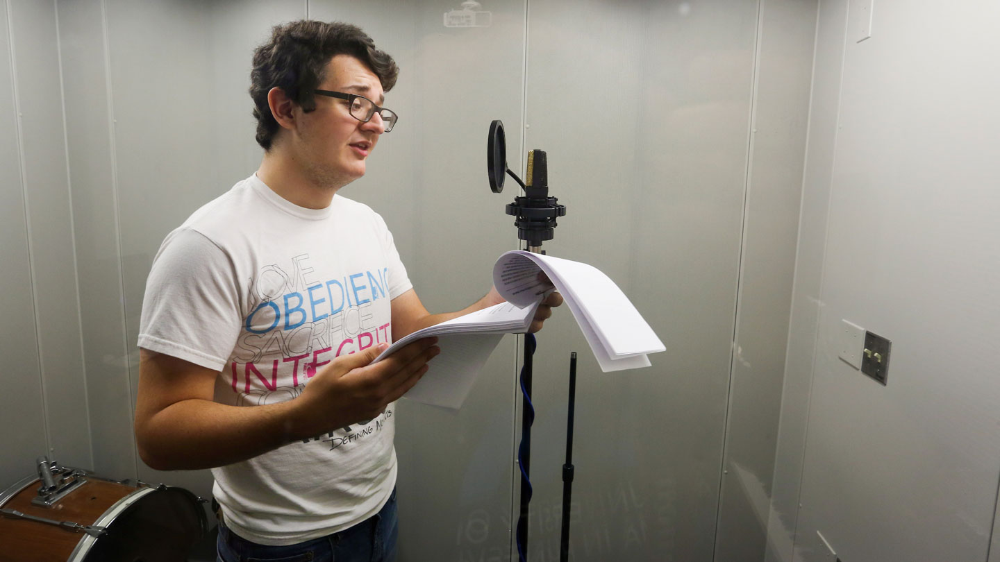 A student speaks from a script into a microphone in a booth