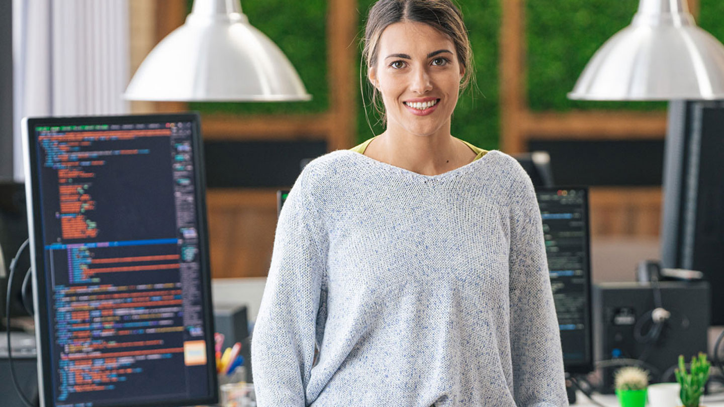A smiling cybersecurity student leans on her desk in front of a vertical screen full of rainbow-colored computer code.