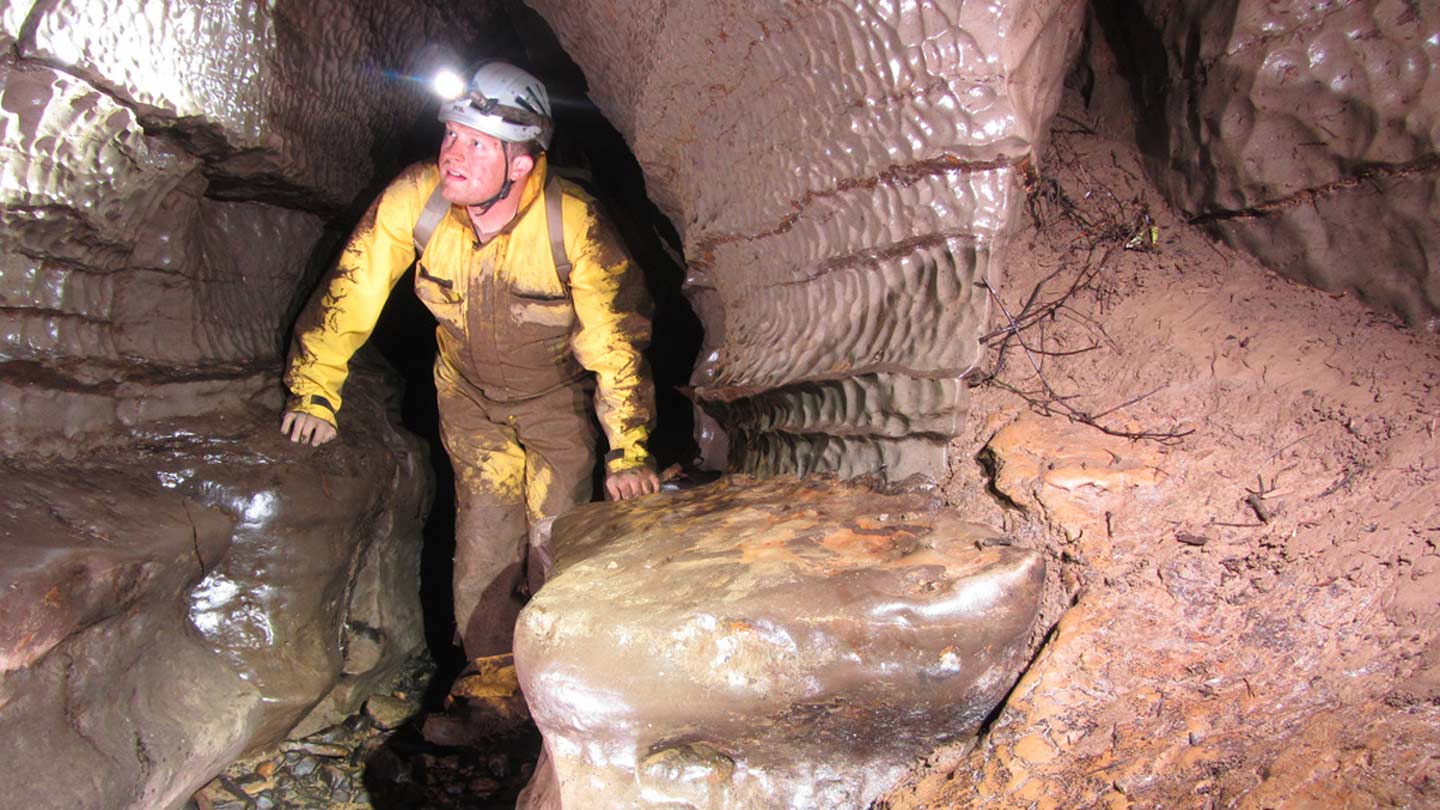 A man with a head lamp covered in mud traverses a cave