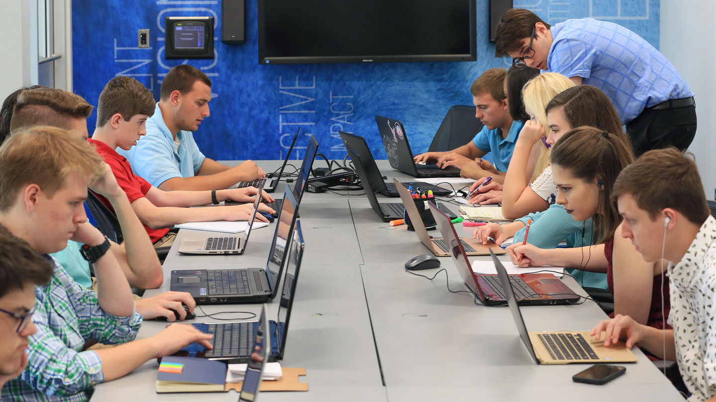 A group of focused students sit at a long table, writing together.
