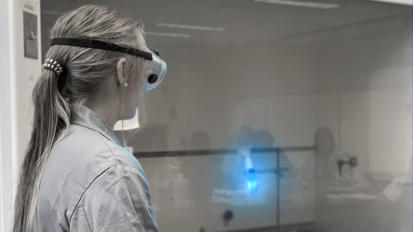 Young woman with a ponytail wearing a lab coat and goggles, facing away to watch an active reaction in a test tube.