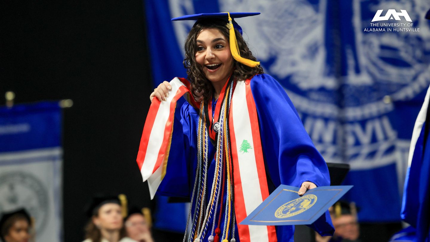 UAH Graduate in blue cap and gown holding diploma, wearing red and white sash with green cedar tree, celebrating at graduation ceremony.