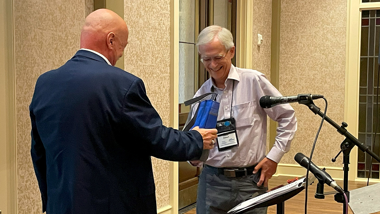 Steve Stewart puts his camera away to receive his Volunteer of the Year award from Board President John Mason at the 2024 Annual Meeting.