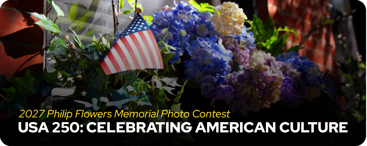 Blue and white flowers plus small American flag underneath window of brick building. Caption is 2027 Philip Flowers Memorial Contest.