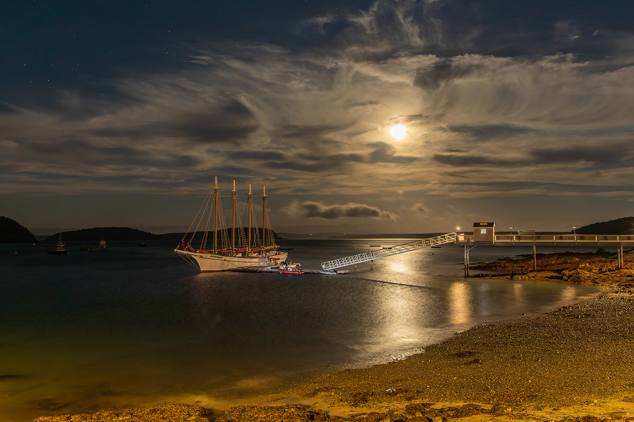 A sailboat docked at the end of long pier at night. The moon is shining through the clouds.