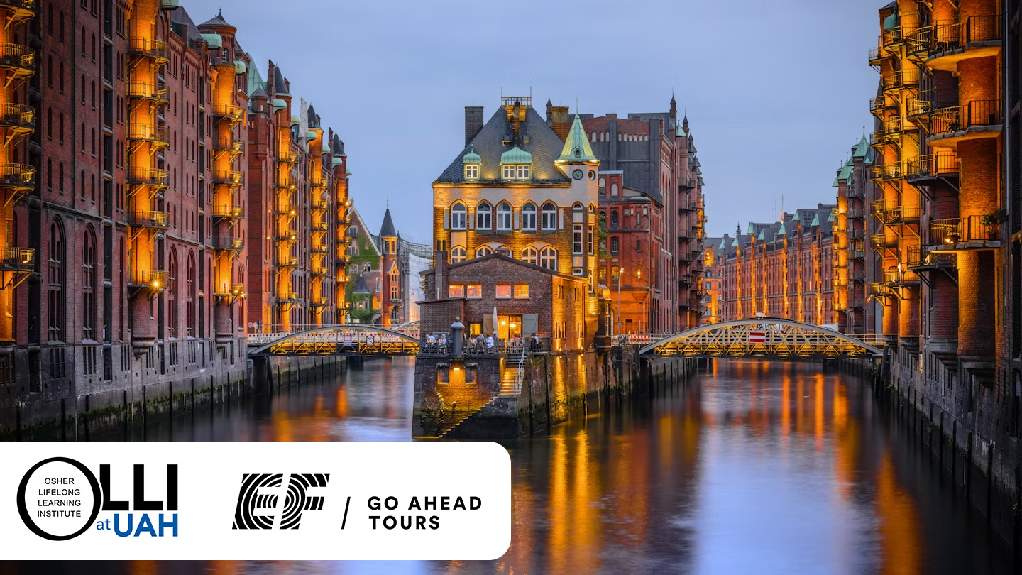 A river running between to streets full of stone buildings. There is a bridge crossing the river with buildings in the middle. It's dusk and all of the buildings lit with warm lights.