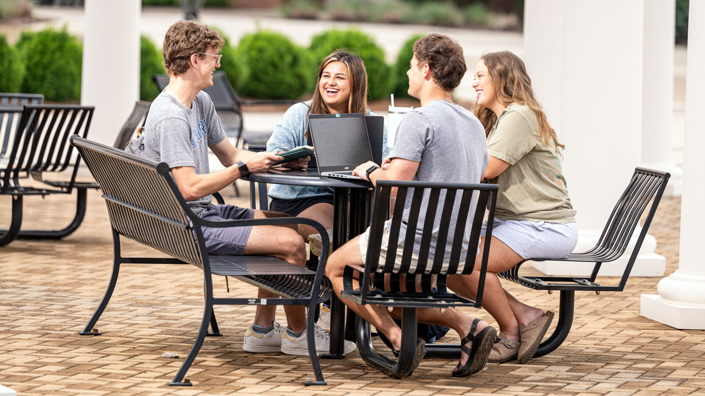 Four UAH students study and laugh together at an outdoor table on the Altenkirk Greenway.