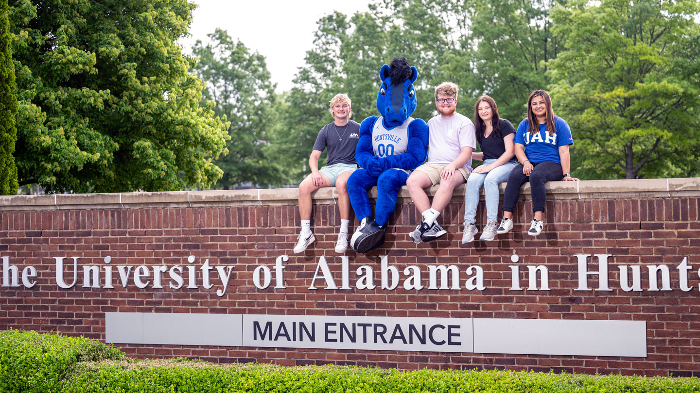 The UAH mascot and students sit on a brick wall at the main entrance to UAH.