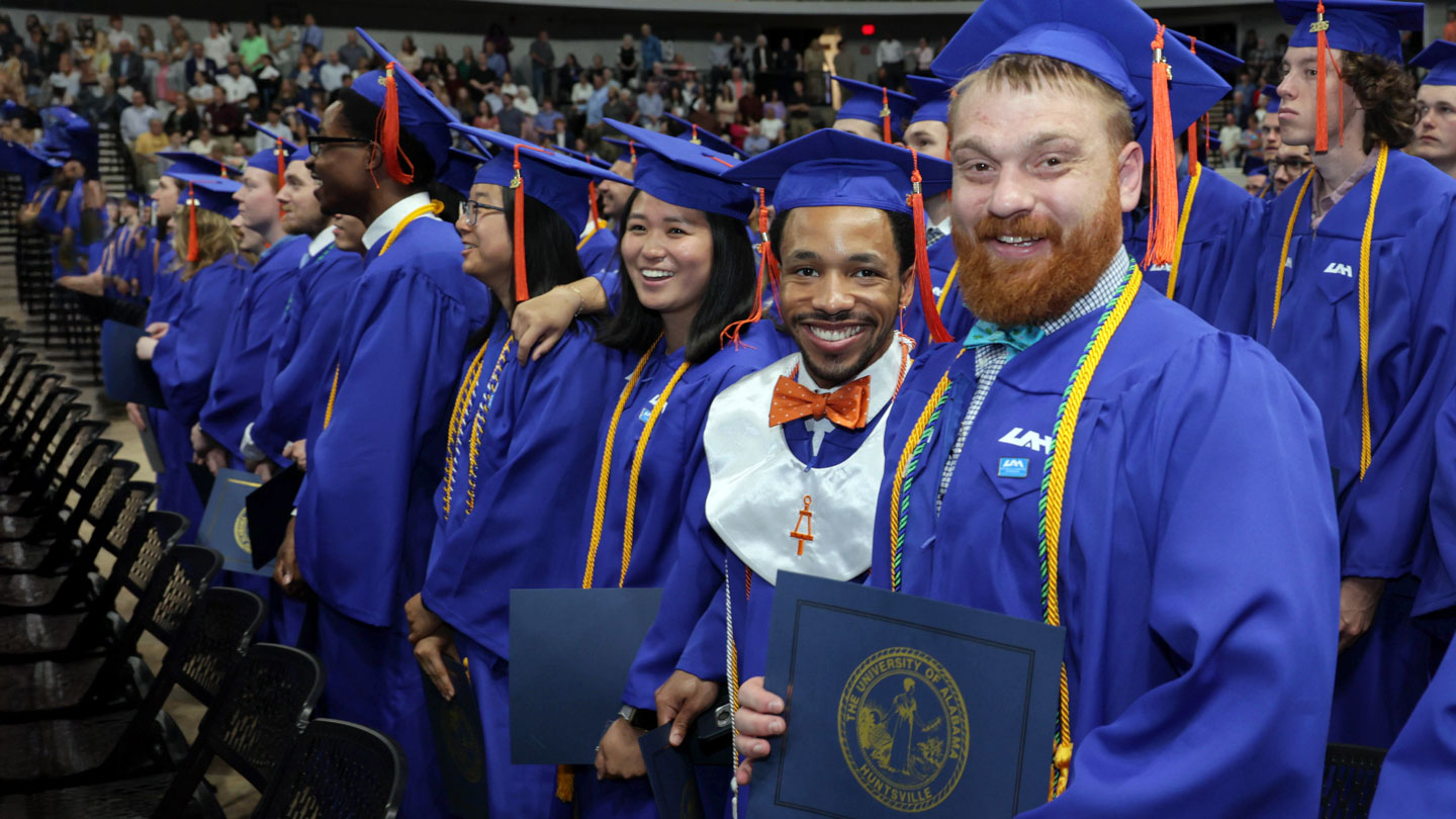 Smiling UAH students in full graduation regalia proudly display their diplomas at  spring commencement in 2026.