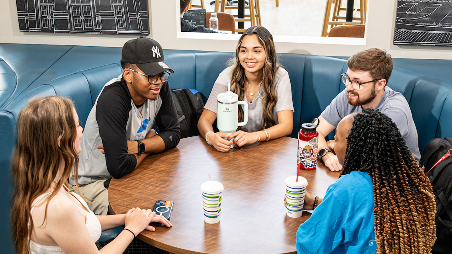 A diverse group of U A H students have a conversation at a round table in the Charger Union building.