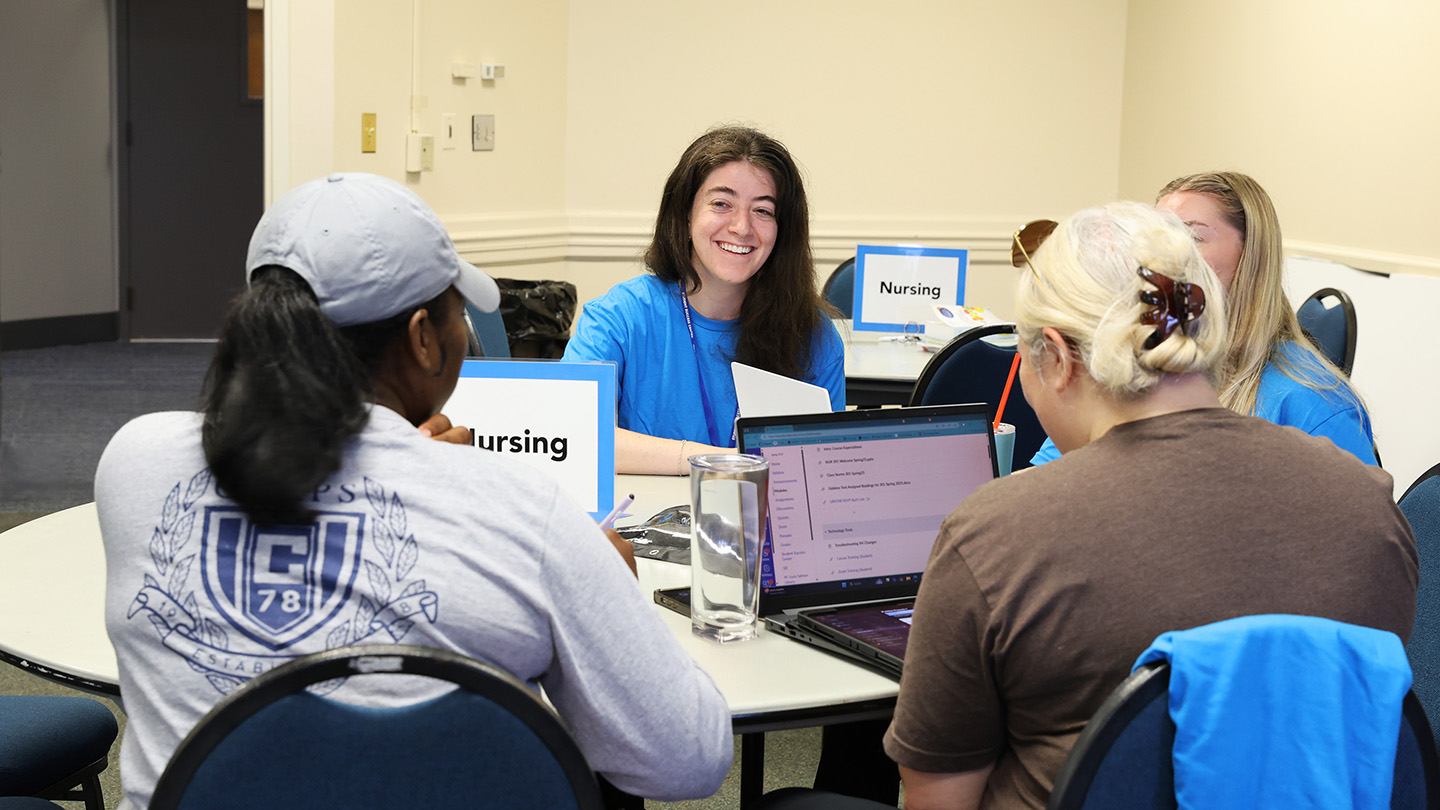 A group of nursing students gathers around a table, collaborating with laptops and materials in a bright classroom setting.