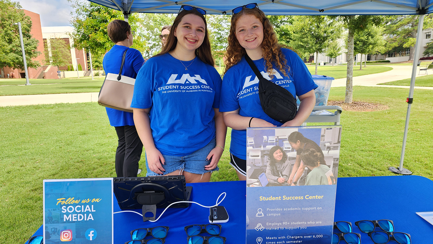 Two UAH young women in blue Student Success Center shirts stand behind a table with information and sunglasses outdoors.
