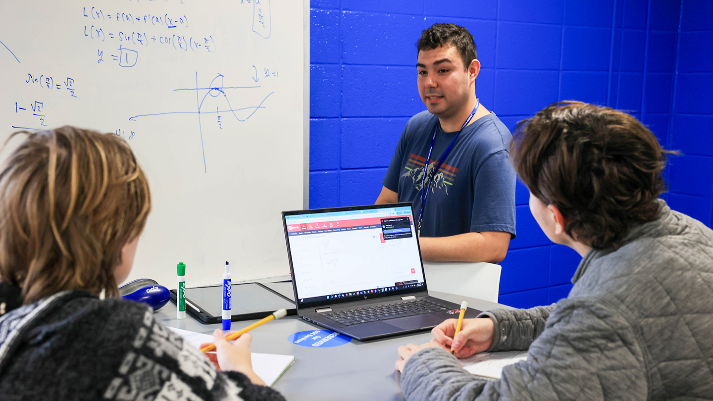 A tutor explains mathematical concepts to two students, with notes and a laptop open on a table against a blue wall.