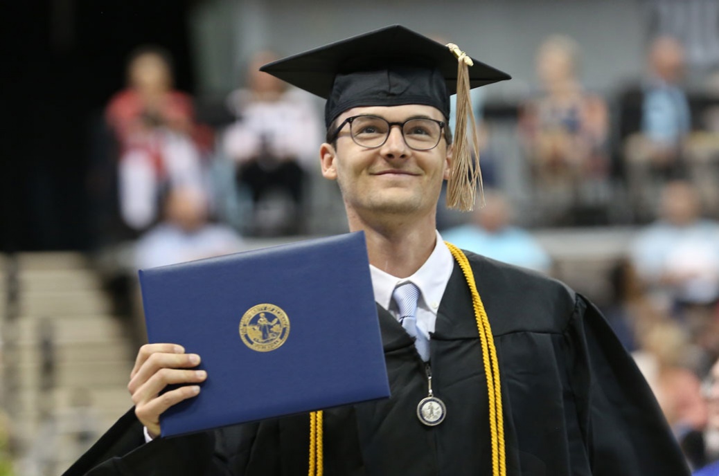 a smiling young man wearing glasses and a graduation robe holds his diploma with a U A H seal on the cover