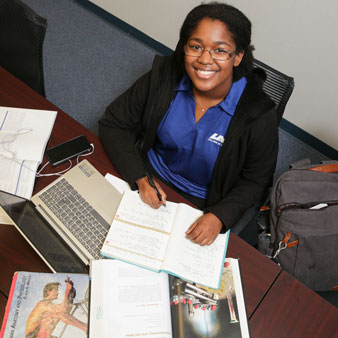 a smiling student studies for exams surrounded my books, notebooks, and a computer