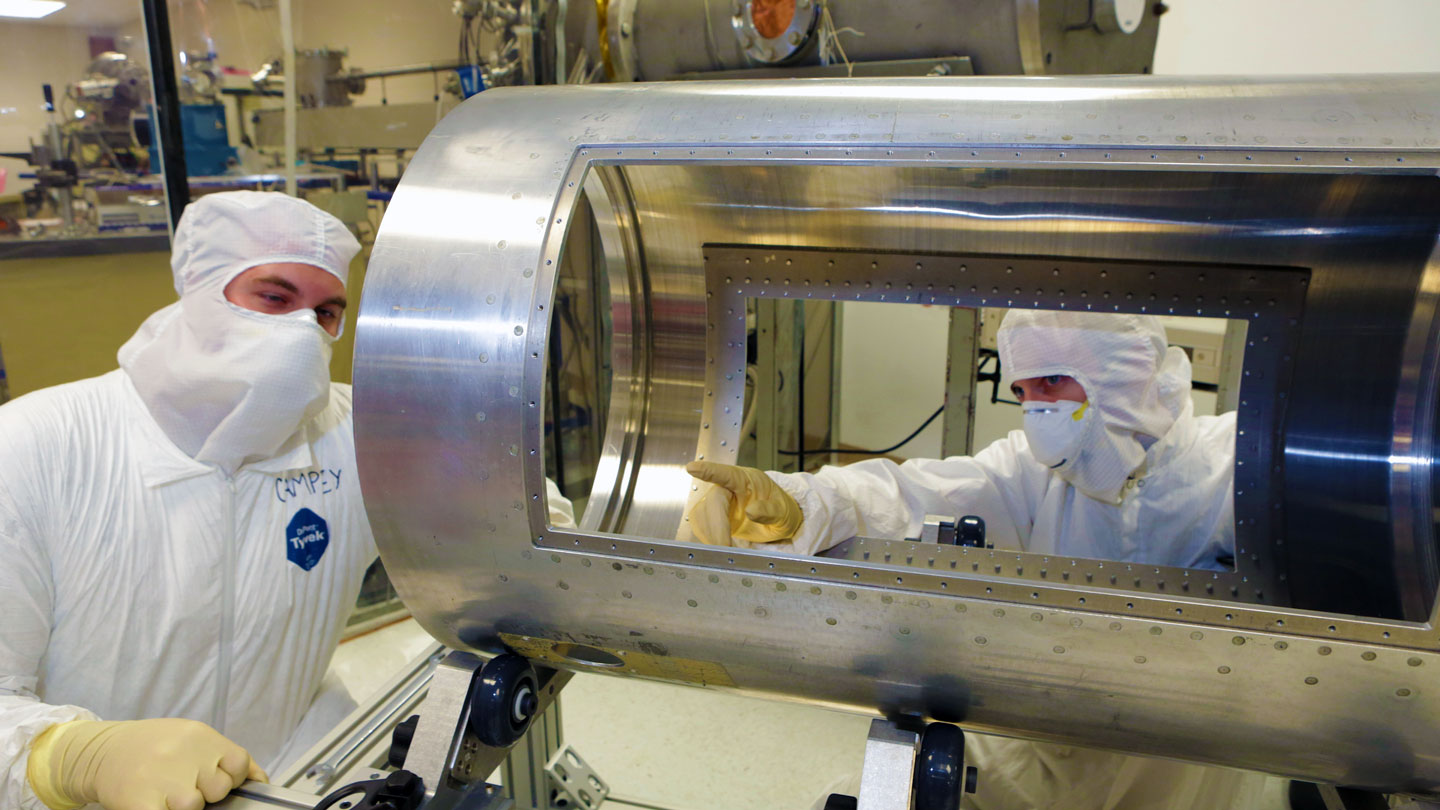Two U A H graduate students dressed in sterile coveralls work on a sensitive piece of equipment in a clean room.