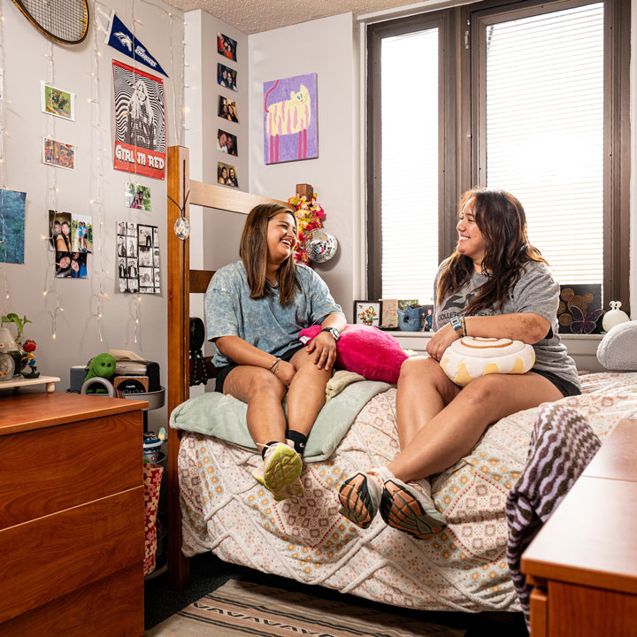 Two students sitting on a dormitory room bed have a conversation.