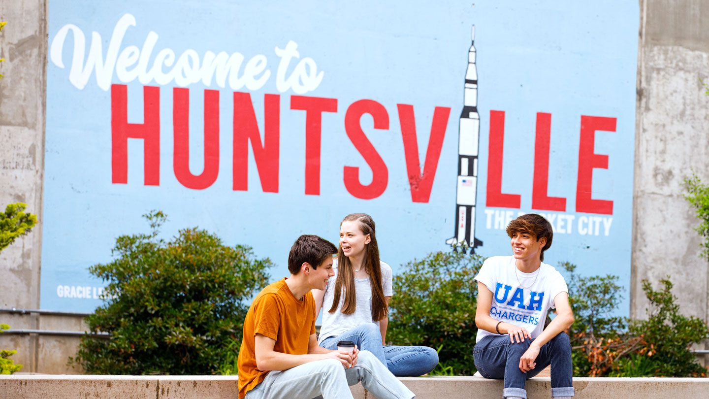 Three smiling U A H students having a conversation in front of a mural of the Saturn V rocket with text that reads Welcome to Huntsville the Rocket City.