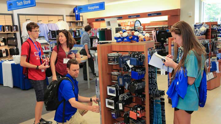 Students browse a store displaying tech and school supplies, with one kneeling to examine products while others look on.