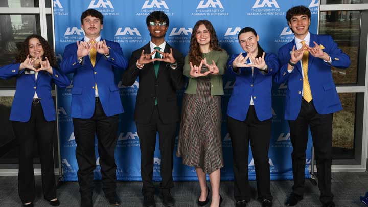 Five individuals in blue blazers pose together in front of a university banner, making hand signs and smiling.