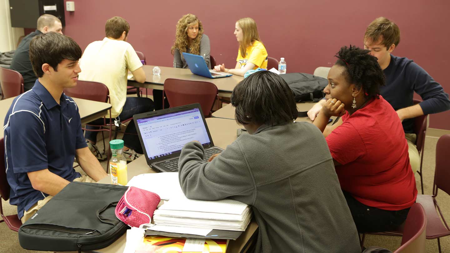 students setting around a table in a classroom with books and computers out, talking.