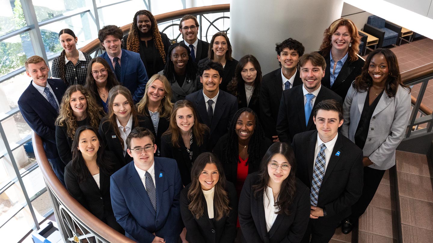 A group of 15 UAH Student Ambassadors in formal attire stands on a staircase in a modern building, with natural light and a curved railing in the background.