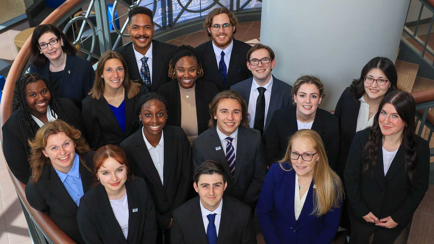 A group of 15 UAH Student Ambassadors in formal attire stands on a staircase in a modern building, with natural light and a curved railing in the background.