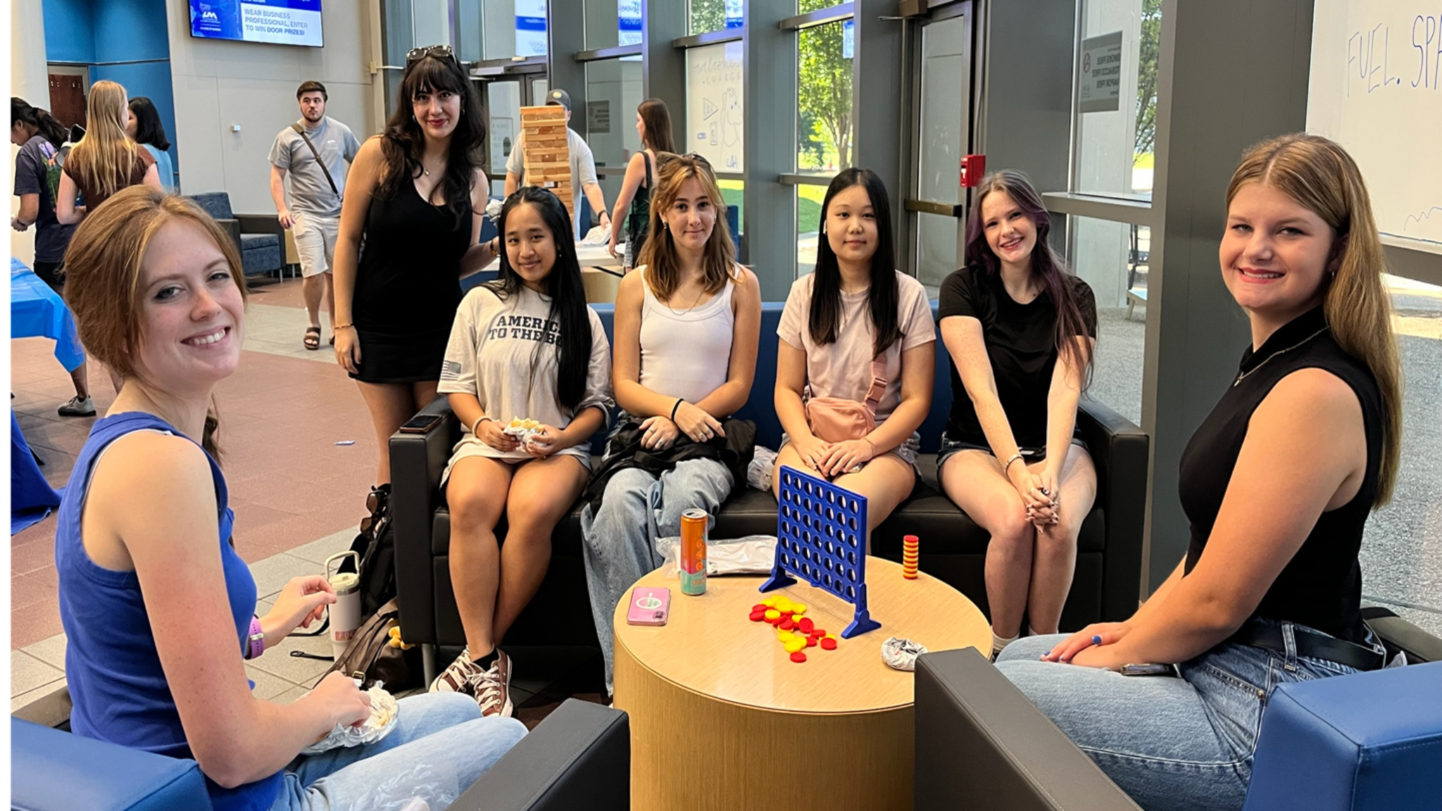 Seven college students gathered in a bright campus atrium, seated on couches and chairs, smiling at the camera with a Connect Four game and drinks on the table.