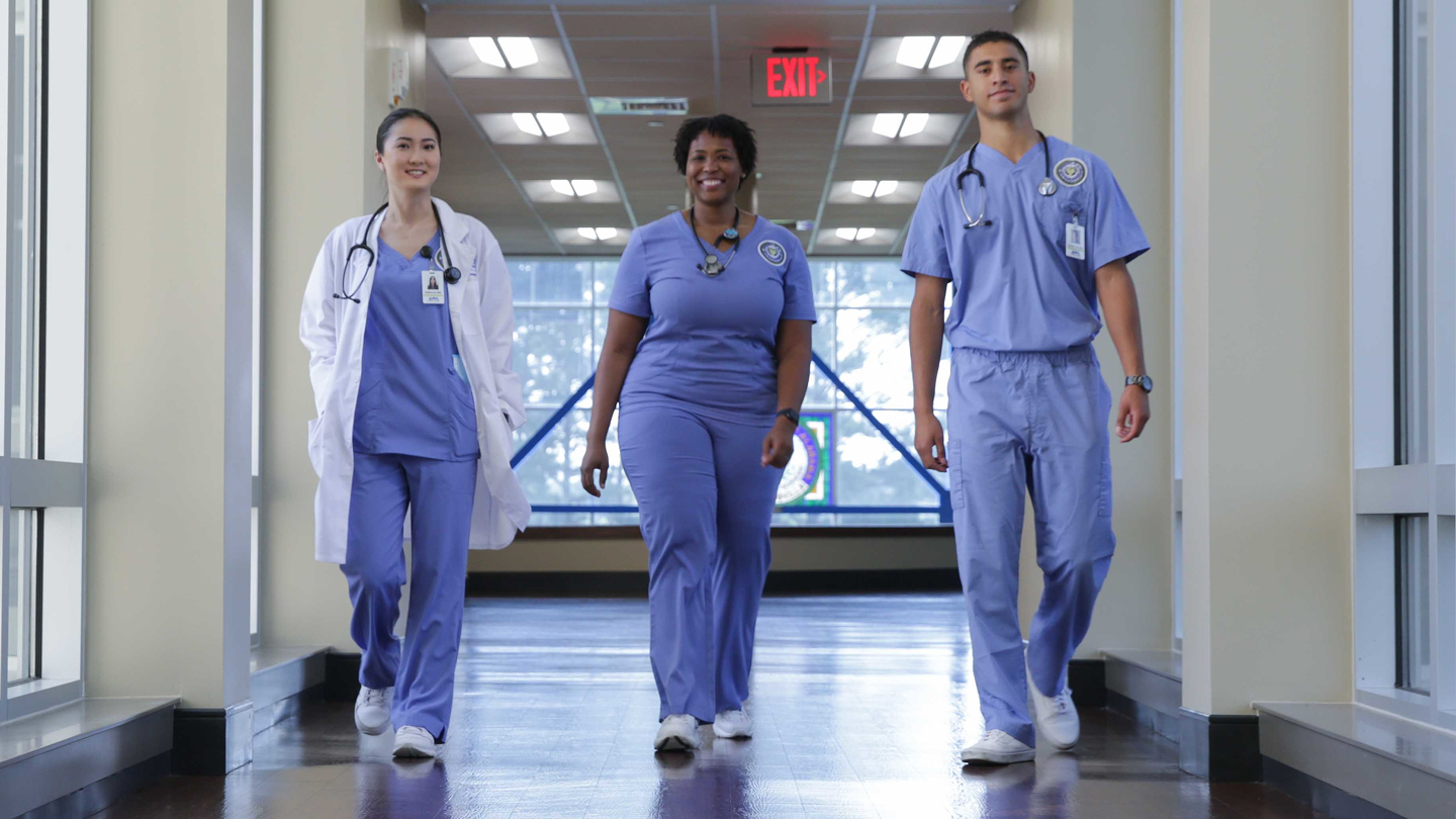 three UAH nursing students in medical uniforms walking down a hospital corridor and confidently smiling