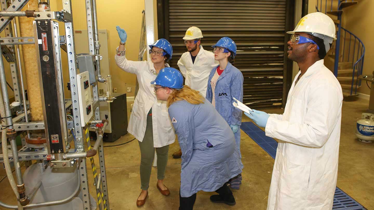 Students standing in lab coats and hard hats looking at machinery.