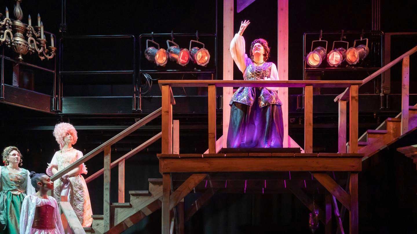A performer in a vibrant purple gown stands on a wooden platform, gesturing dramatically, surrounded by fellow actors in costumes.