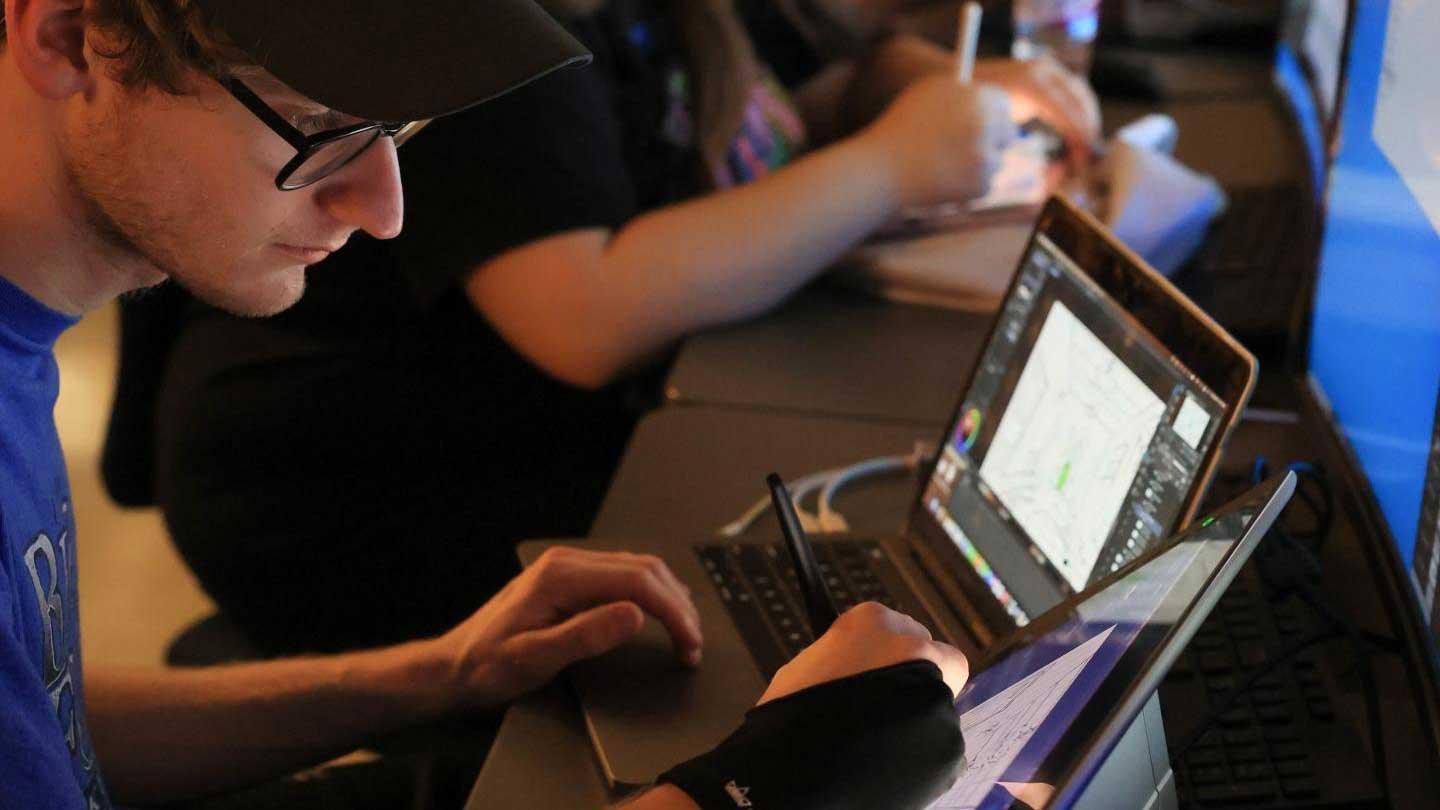 A person using a digital tablet and stylus while seated at a desk, with art software open on a laptop screen nearby.