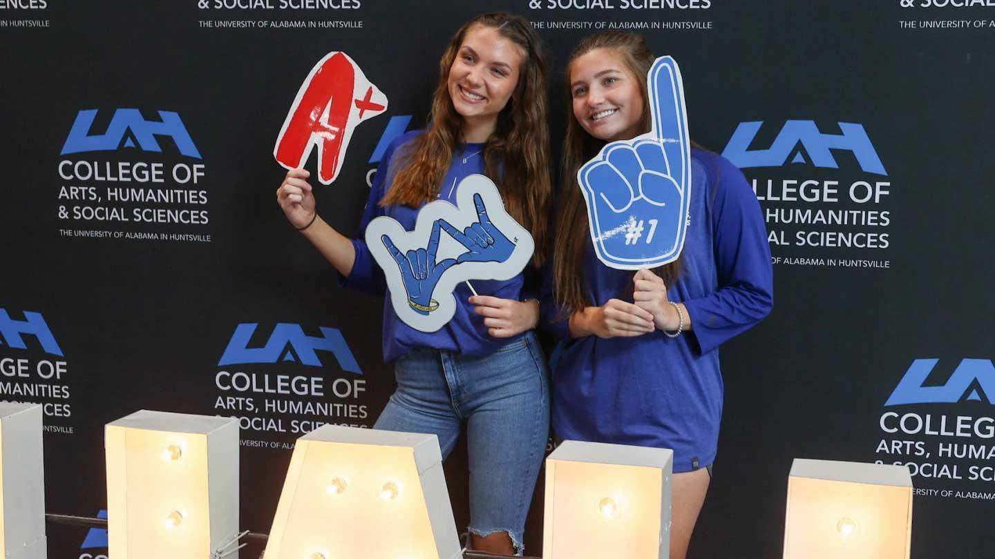 Two individuals hold colorful foam hand signs promoting school spirit, standing in front of a university of Alabama in Huntsville backdrop, dressed in blue shirts.