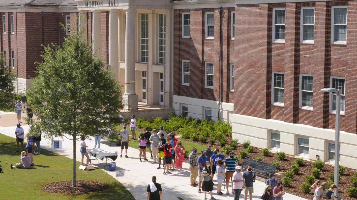 A crowd of students gathers outside Morton Hall, with some waiting in line and others sitting on the grass under sunny skies.