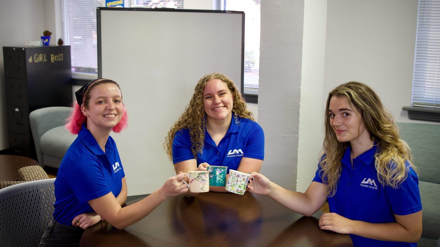 three uah honors advisors clink coffee mugs happily with a white board and girl boss sign in the background