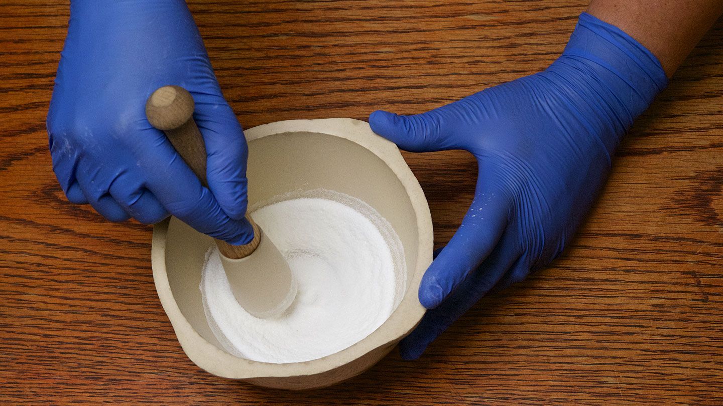 A UAH student's hands wearing blue gloves crushing medication with a mortar and pestle on a wood table