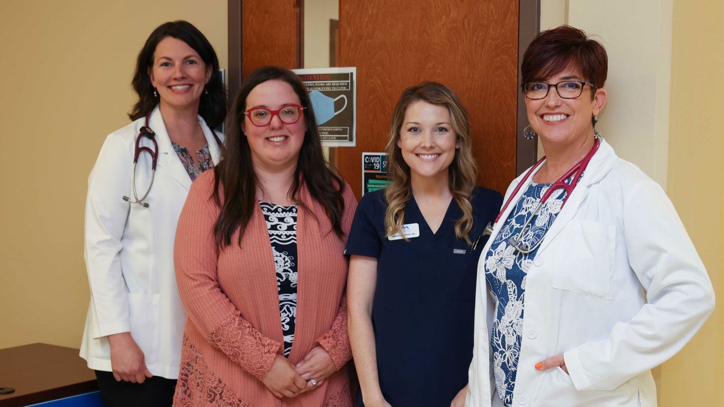 four women stand in a doctors office, two in lab coats, one in scrubs, and one in plain clothing