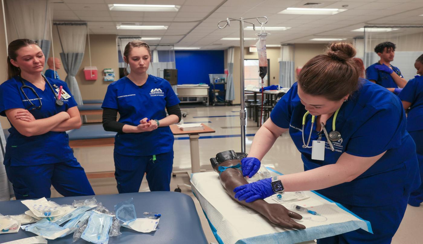 A group of students practicing IV insertion techniques using a task trainer arm. ABSN-NUR-14-