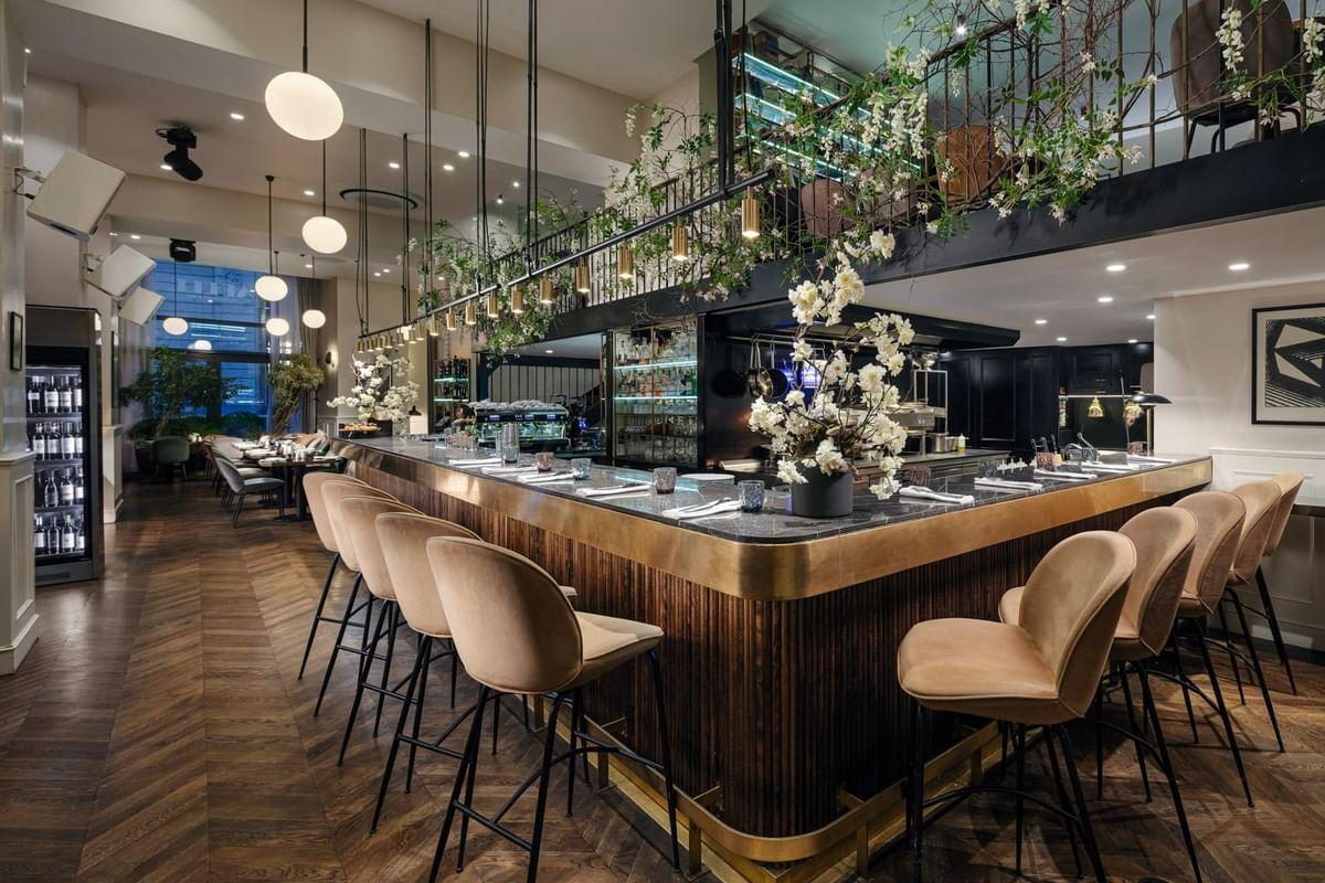 Modern restaurant (Adele) bar with brass counter, tan velvet stools, and a plant-covered mezzanine above.