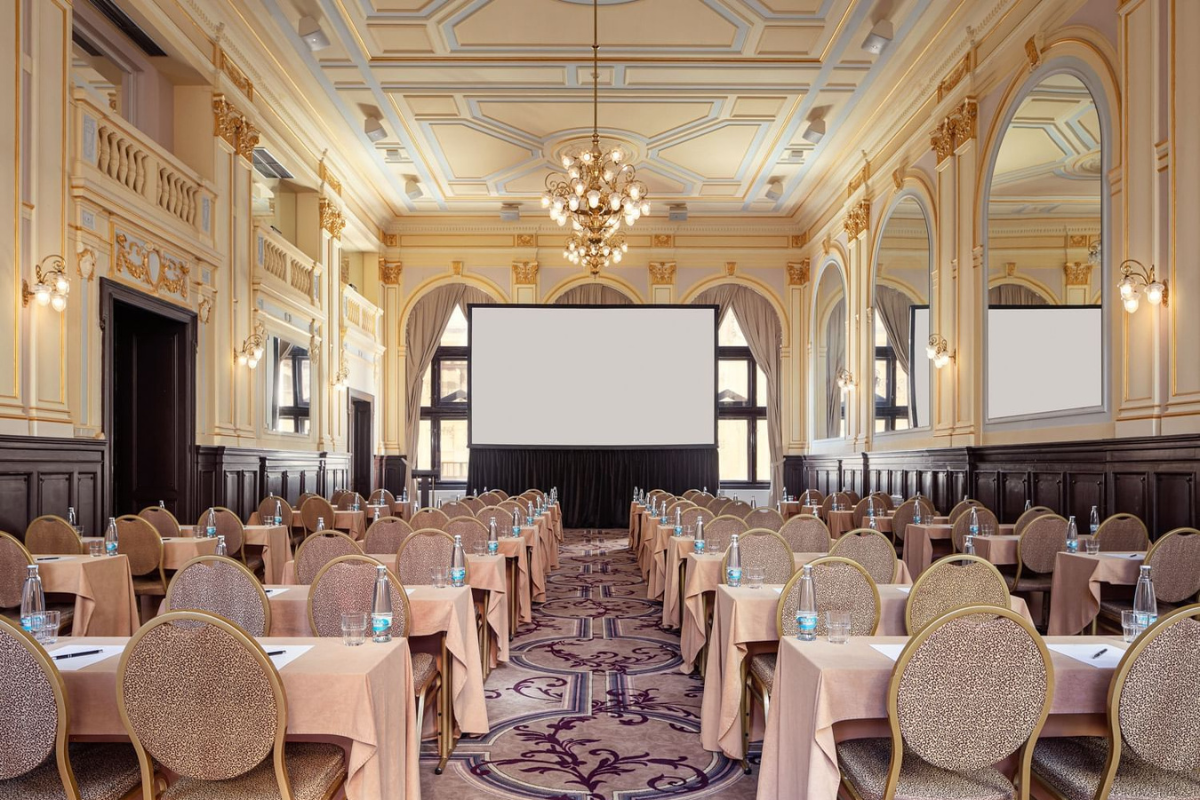 Ornate gilded ballroom set classroom-style with rows of draped tables facing a projector screen.