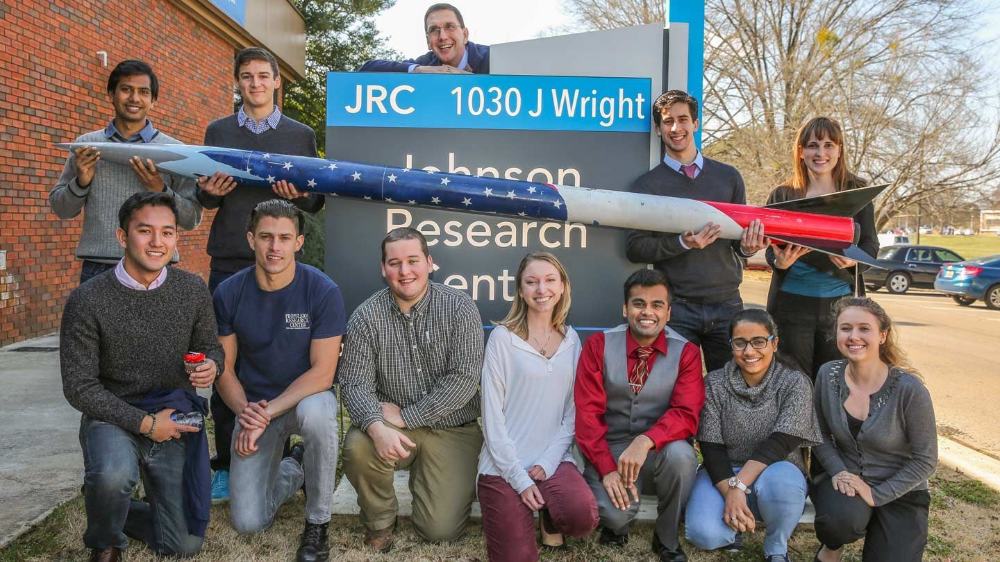 students hold a small rocket in front of the jrc building sign