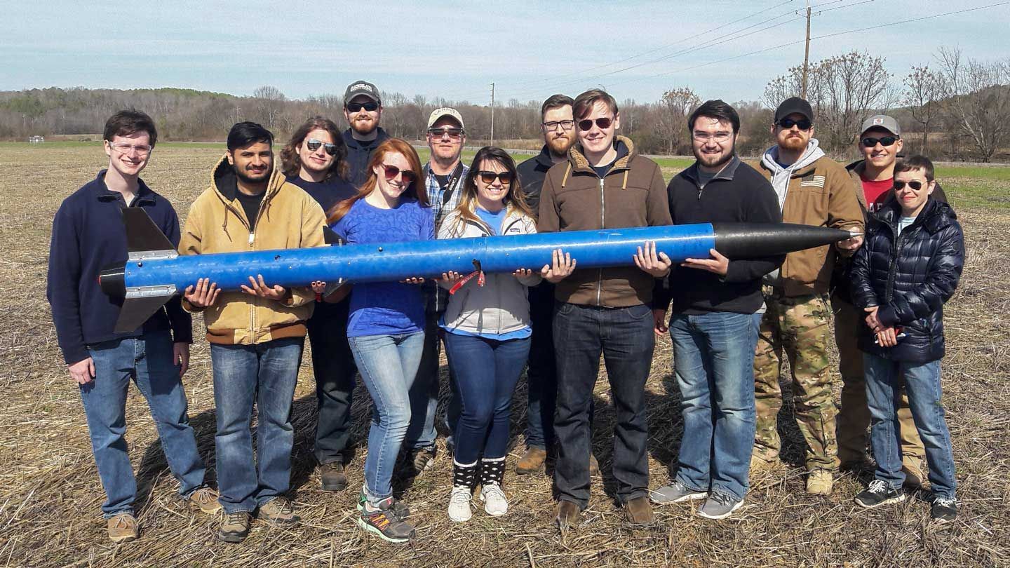 Students proudly hold a small rocket in a field